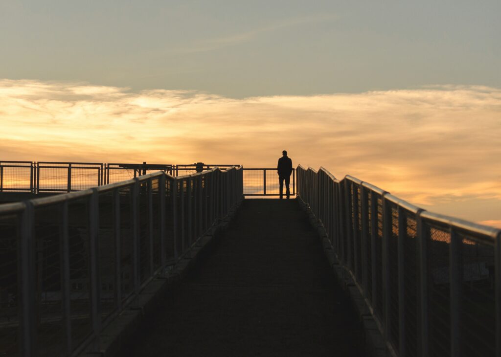 Silhouette of a solitary veteran standing on a bridge walkway at sunset, with orange and yellow clouds in the background.