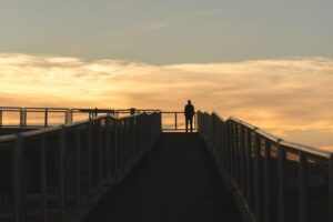 Silhouette of a solitary veteran standing on a bridge walkway at sunset, with orange and yellow clouds in the background.