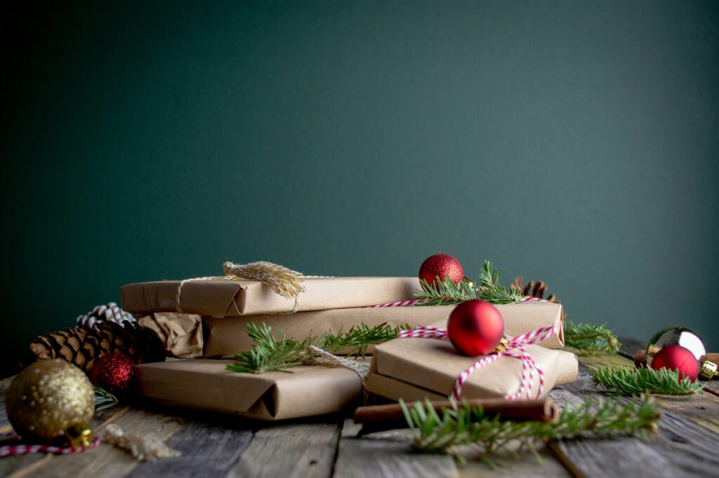 Wrapped Christmas gifts with red ornaments and pine branches rest on a wooden surface against a dark green background.