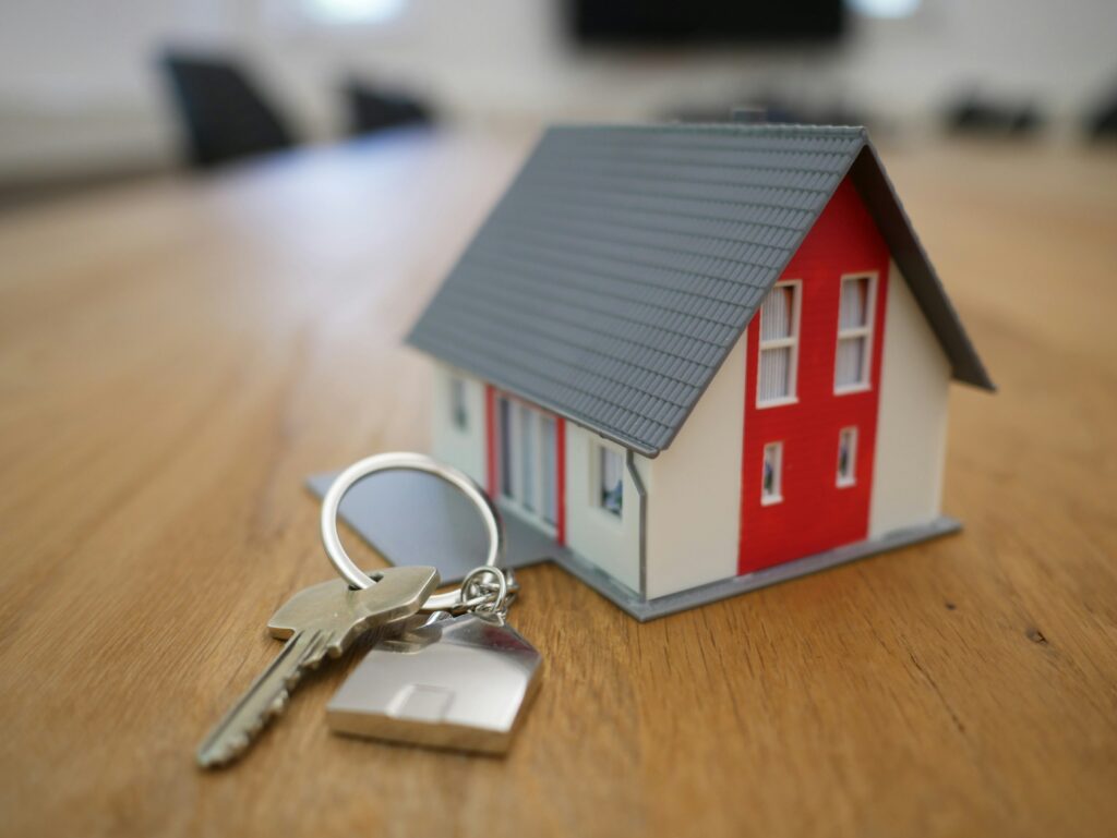 A small model house with a red facade rests on a wooden table next to a set of keys, symbolizing the journey towards securing a mortgage.