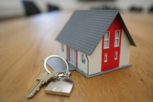 A small model house with a red facade rests on a wooden table next to a set of keys, symbolizing the journey towards securing a mortgage.