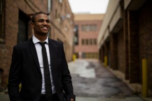 A person in a black suit and tie stands smiling in an urban alleyway with brick buildings, celebrating the vibrant diversity that surrounds them.