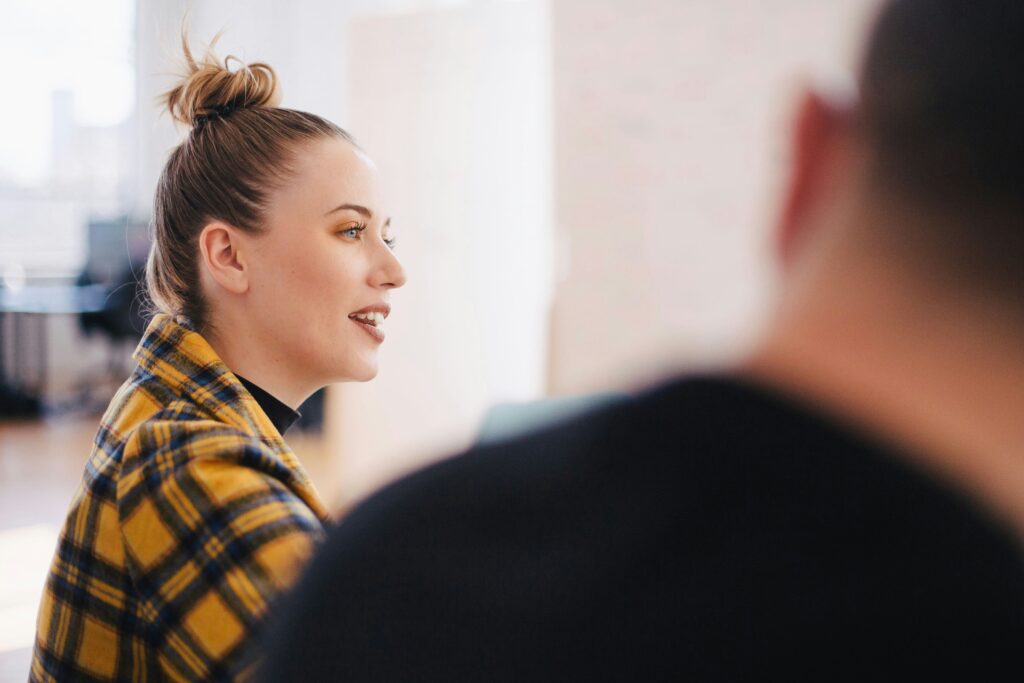 A woman in a yellow plaid shirt is speaking confidently in a bright room, reminiscent of Kamala Harris's poised demeanor. Another person's head is blurred in the foreground.