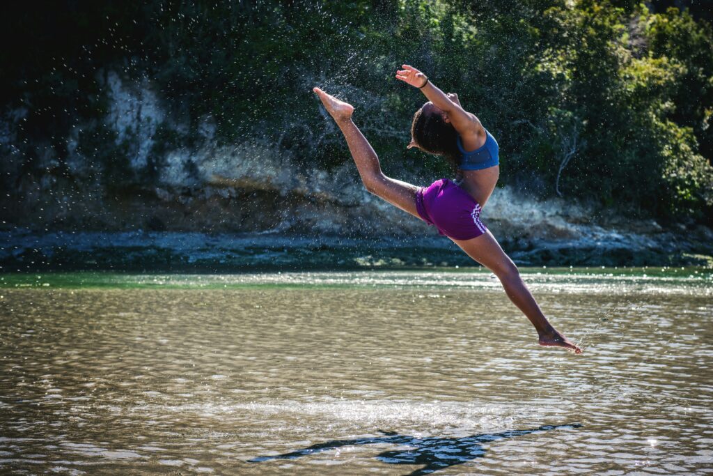 Dressed in athletic attire, the person performs a high leap over shallow water, capturing the essence of movement with one leg extended and arms outstretched. Droplets of water hang suspended in the air against a backdrop of trees and greenery.