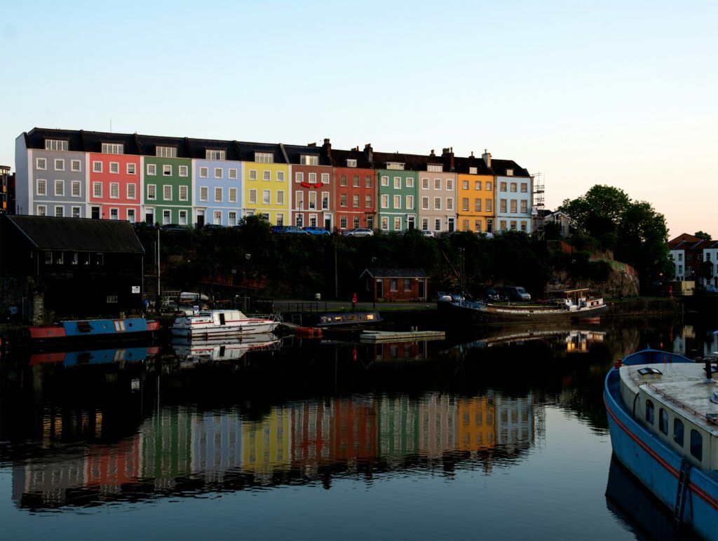 A row of colorful houses reminiscent of charming UK areas is reflected in the water at sunset, with boats docked along the canal.