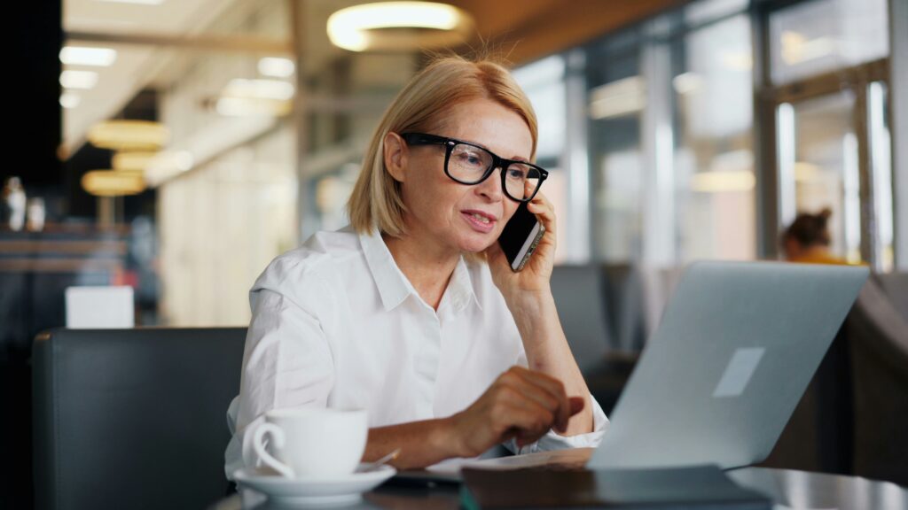 An executive-level woman with glasses is sitting at a table, efficiently multitasking between her phone and laptop. A cup rests beside her, reflecting her busy schedule.