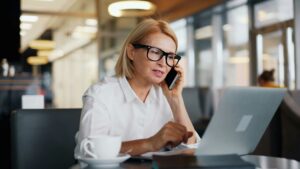 An executive-level woman with glasses is sitting at a table, efficiently multitasking between her phone and laptop. A cup rests beside her, reflecting her busy schedule.