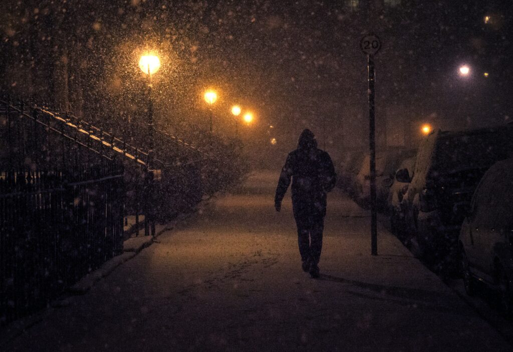 A person strolls down a snow-covered street during the darker months, the path illuminated by streetlights, with cars parked on the right.