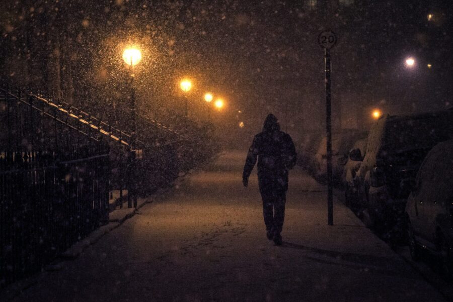 A person strolls down a snow-covered street during the darker months, the path illuminated by streetlights, with cars parked on the right.