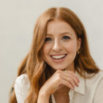 Dr. Emily, with long red hair, wearing a white blouse and blue jeans, sits on a chair and smiles at the camera.