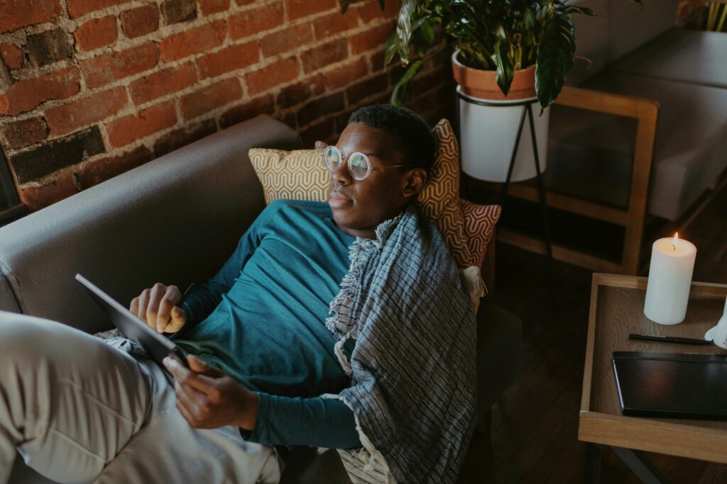 A person wearing glasses, wrapped in a blanket, quietly quits the chaos of the day as they relax on a couch with a tablet. Nearby, a lit candle and a potted plant create a soothing atmosphere.