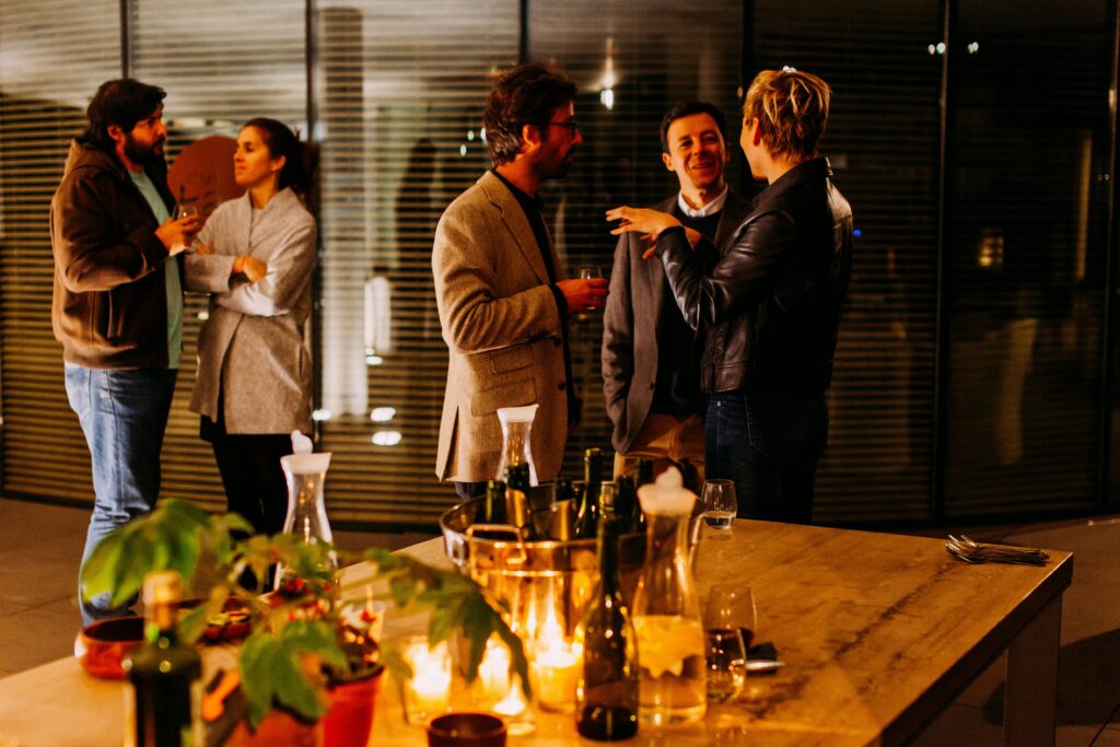A group of people stand and chat at a dimly lit Christmas party, with a table holding bottles, glasses, and candles in the foreground.
