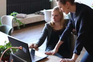 Two women in an office setting embody compassionate leadership as they collaborate over a laptop screen, with one seated and the other standing, fostering a supportive and engaging work environment.