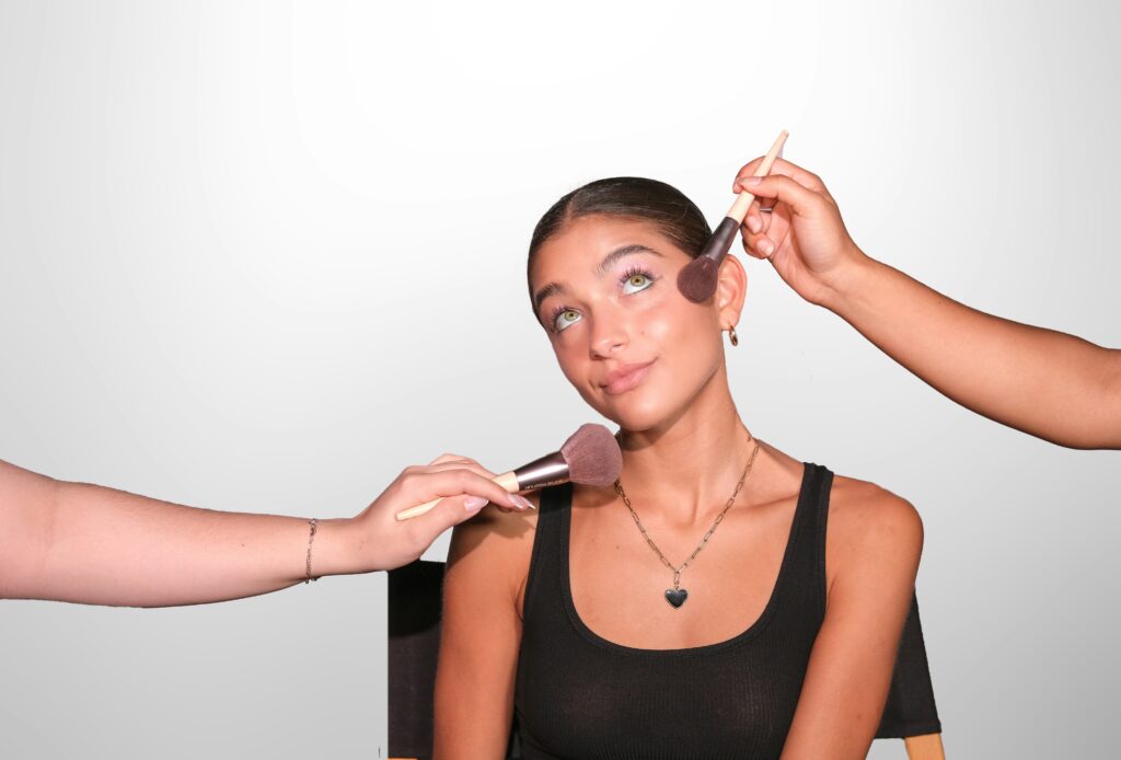 Person sitting in a chair, wearing a black tank top, skillfully applying makeup with brushes to their face against a plain background, perhaps preparing for another day of tackling tasks alongside those occasionally annoying co-workers.
