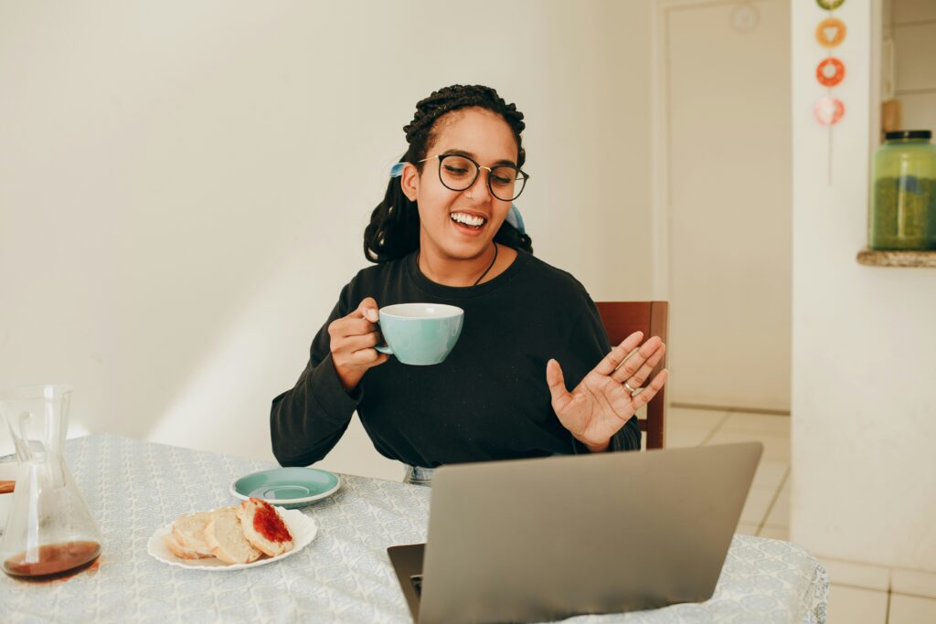 A person smiles during an online call on a laptop, holding a mug. Nearby, slices of bread with jam are on the table.