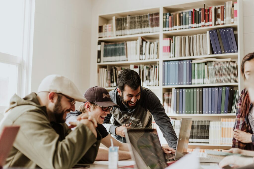 A group of people smiling and collaborating around a laptop in a library setting, blending the charm of bookshelves with the camaraderie of working from the office.