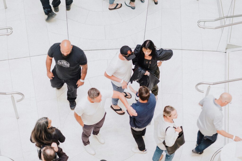 A group of vibrant personalities interacts in a busy, white-tiled space with stairs. Some are standing in animated conversation, while others are walking briskly.