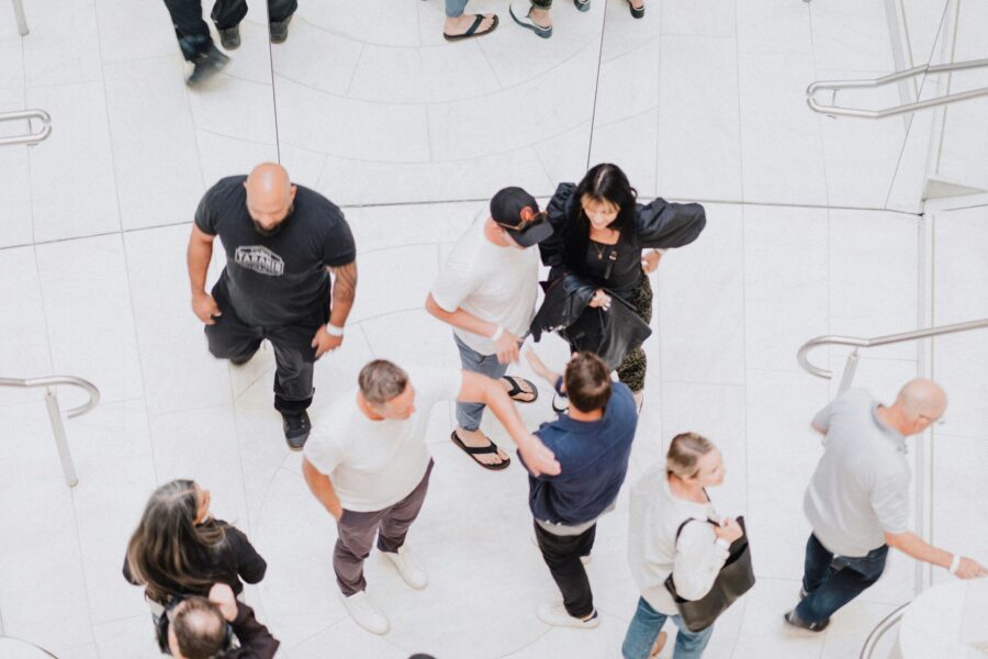 A group of vibrant personalities interacts in a busy, white-tiled space with stairs. Some are standing in animated conversation, while others are walking briskly.