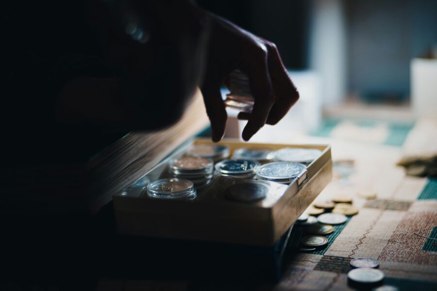 A hand selects a coin from a box containing various coins on a table, with scattered coins around. The scene is dimly lit, symbolizing the elusive struggle against the gender pay gap and creating a focused atmosphere.