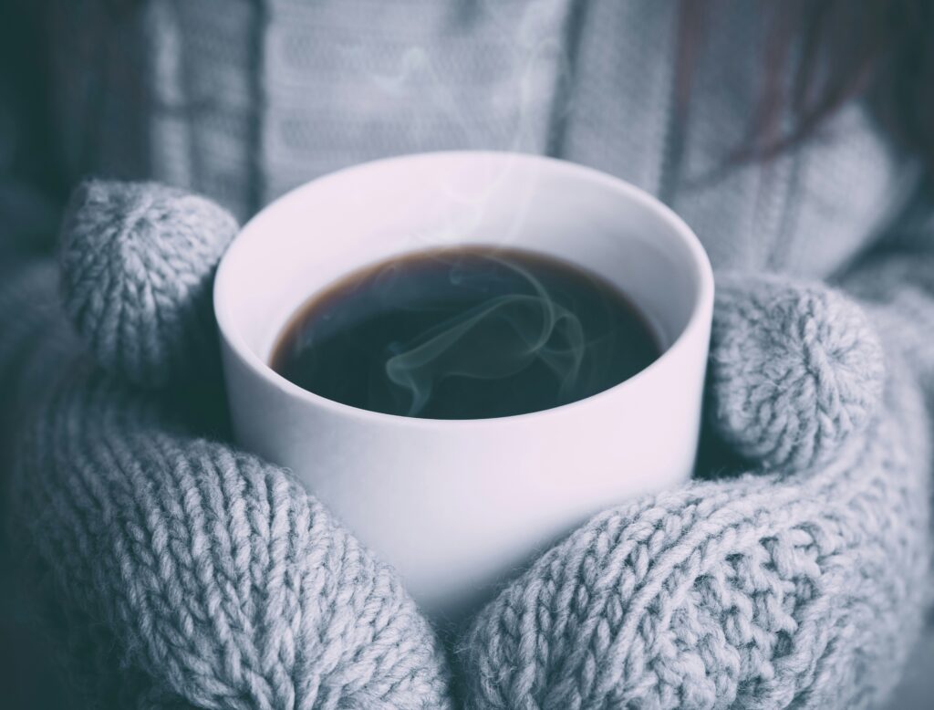 A person wearing gray knitted mittens holds a steaming cup of coffee, savoring the warmth that not only comforts but also invigorates their immune system.