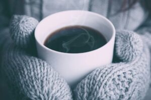 A person wearing gray knitted mittens holds a steaming cup of coffee, savoring the warmth that not only comforts but also invigorates their immune system.