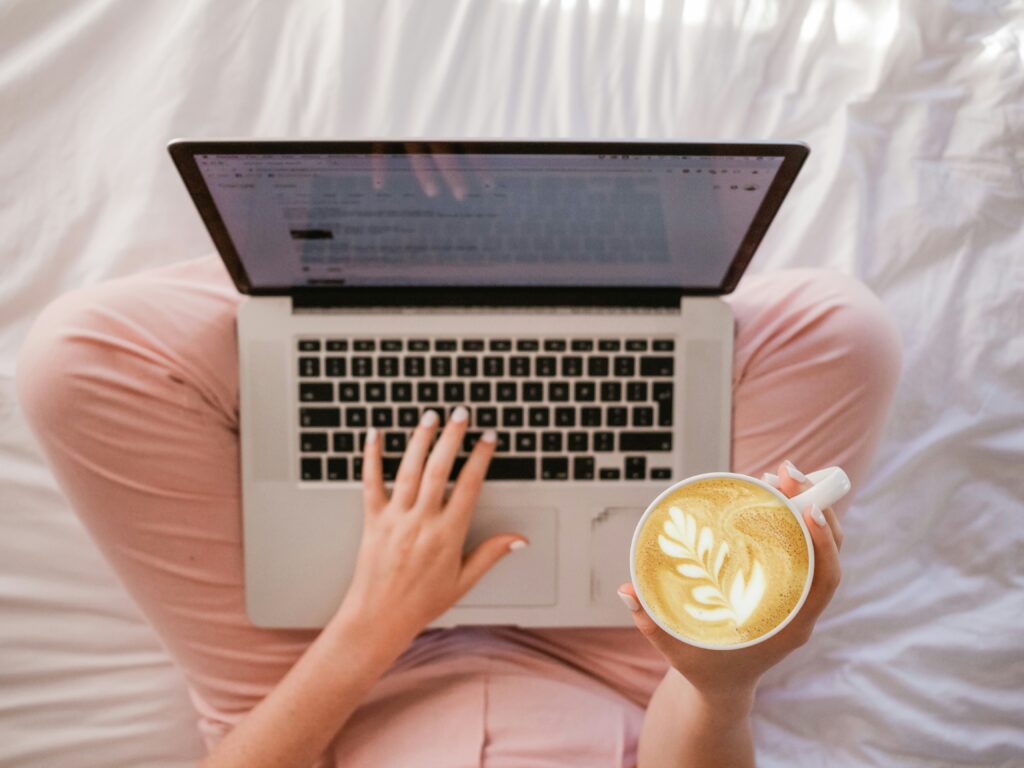 A person in pink pants is on a wockcation, effortlessly balancing work and relaxation. They're using a laptop on their lap while savoring a latte with leaf art in one hand, all from the comfort of a white bed.