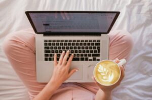 A person in pink pants is on a wockcation, effortlessly balancing work and relaxation. They're using a laptop on their lap while savoring a latte with leaf art in one hand, all from the comfort of a white bed.