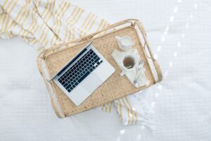 A laptop, cup of tea with creamer, and a spoon rest on a wicker tray atop a white and yellow striped blanket, creating a cozy setup that complements workplace health by offering moments of relaxation in your busy day.