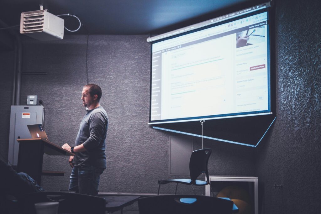 A man stands at a podium giving a presentation in a dimly lit room, with a screen displaying a webpage behind him. On the frontline of digital innovation, he captivates his audience with insights that illuminate the path forward.