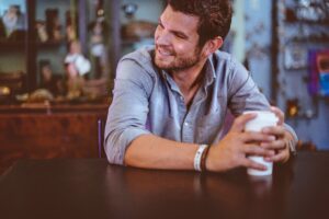 A man in a gray shirt holds a coffee cup, sitting at a table and smiling, embodying the essence of men's mental health. The background reveals shelves adorned with various items, adding warmth to the tranquil scene.