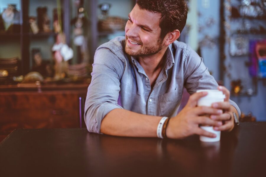 A man in a gray shirt holds a coffee cup, sitting at a table and smiling, embodying the essence of men's mental health. The background reveals shelves adorned with various items, adding warmth to the tranquil scene.