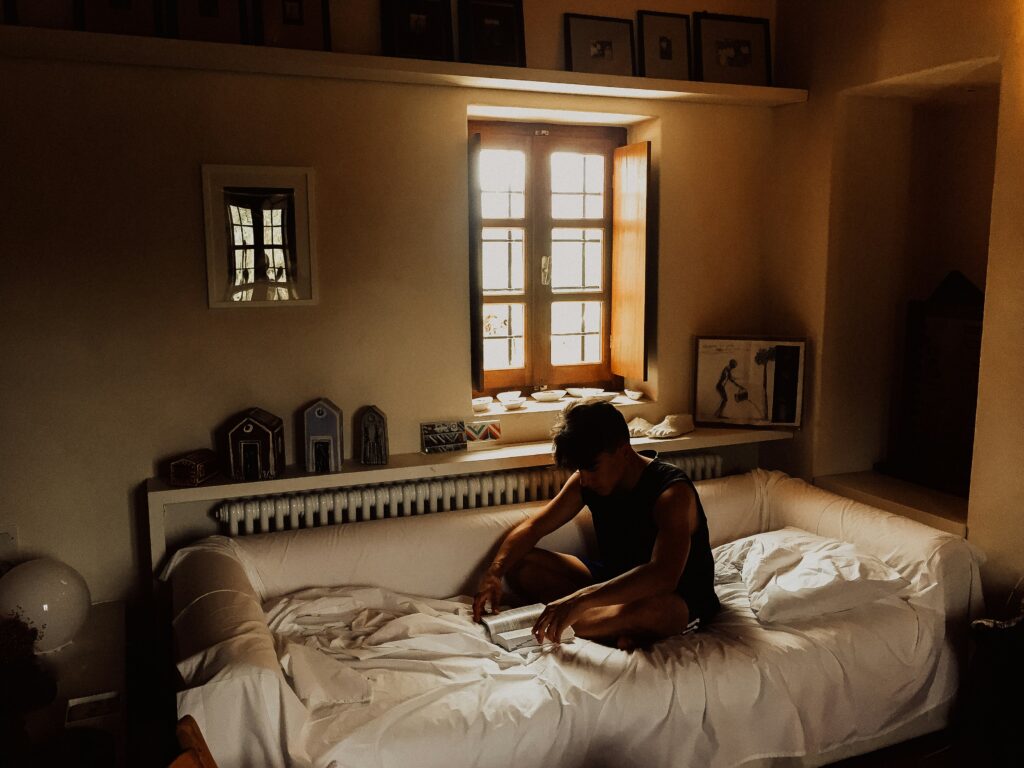 A person sits reading on a white sofa in a dimly lit room, nurturing their mental health while surrounded by a window and framed pictures on the walls.