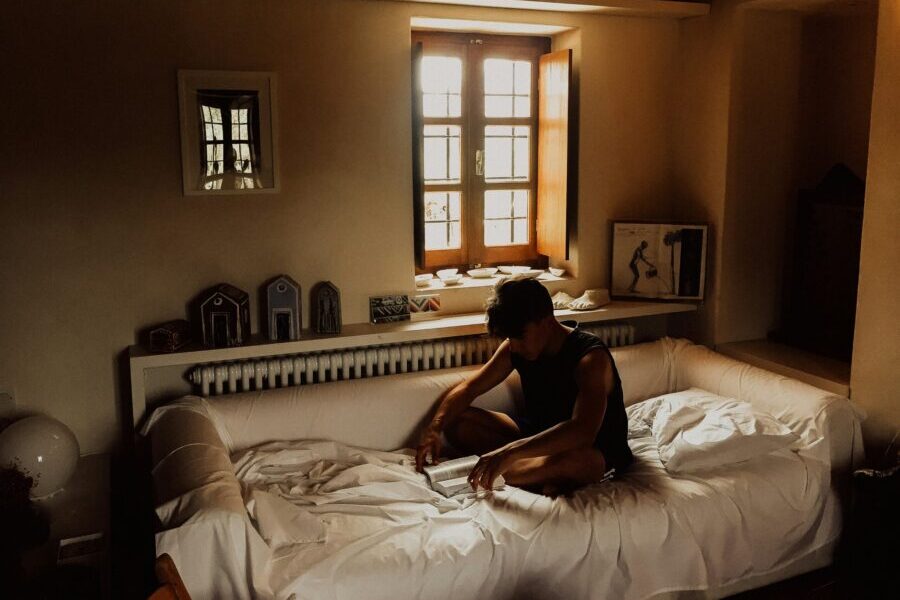 A person sits reading on a white sofa in a dimly lit room, nurturing their mental health while surrounded by a window and framed pictures on the walls.