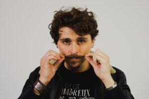 A man with curly hair holds his mustache with both hands, showcasing a playful nod to the importance of men's health and grooming.