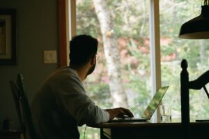 A person engaged in remote work sits at a desk with a laptop, occasionally gazing out the window.