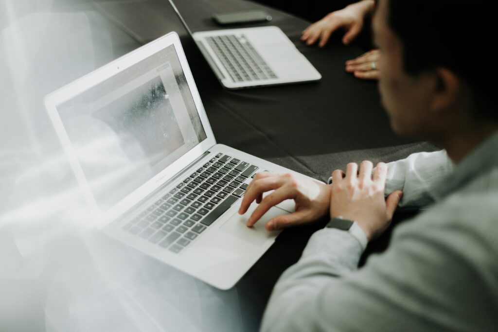An HR leader intensely focuses on their laptop screen at a desk, while another laptop and a pair of hands are visible in the background, signaling a collaborative environment.