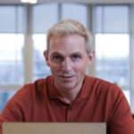 Matthew Norbury, with short light hair, sits at a desk in his red shirt. The office setting is enhanced by natural light and plants as he works diligently on his laptop.