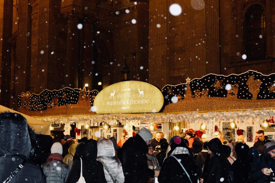 A festive crowd gathers at a lively market with snow falling, under a sign that reads 