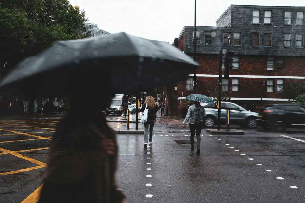 People with umbrellas, braving a rainy street at an intersection, move through a scene that could have inspired an album cover for SAD. They're surrounded by buildings and the gentle chaos of moving cars, each step echoing the melancholy rhythm of the rain.
