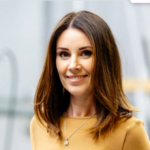 Rebecca Sutherland, a smiling woman with shoulder-length brown hair, wears a mustard-colored top and a necklace as she stands against a blurred background.