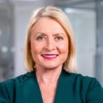 Sheila Flavell, a confident woman with blonde hair, stands with crossed arms in a modern office setting, radiating poise in her dark green blazer.