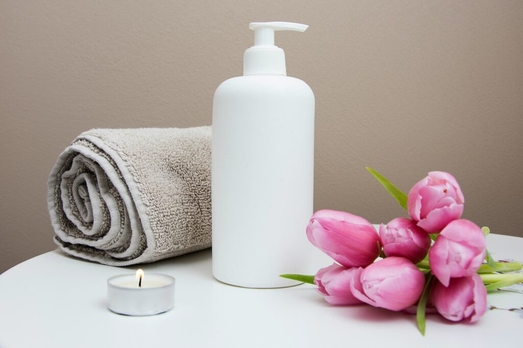 A white lotion bottle sits beside a rolled towel, pink tulips, and a lit tea candle on a white surface, creating a serene scene inspired by the wellness industry.