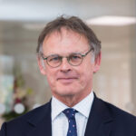 Stewart Sharman, a man in a suit and tie, stands confidently in an office with glass walls.