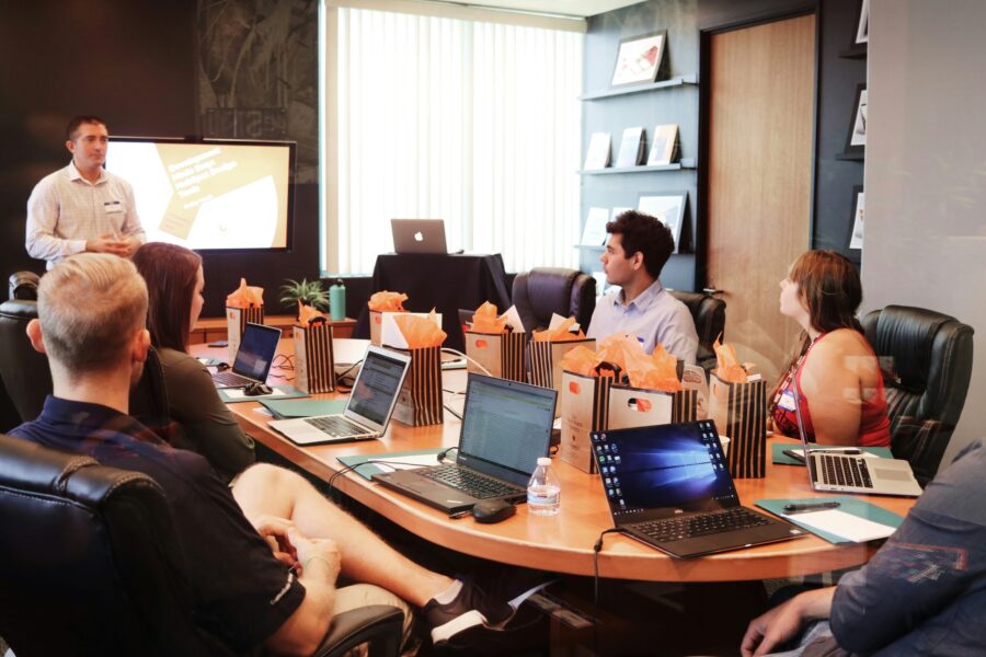 A group of people in a conference room, seated around a table with laptops, listens intently to a presentation focused on wellbeing. A standing speaker delivers insights as a screen displays slides filled with text and graphics.