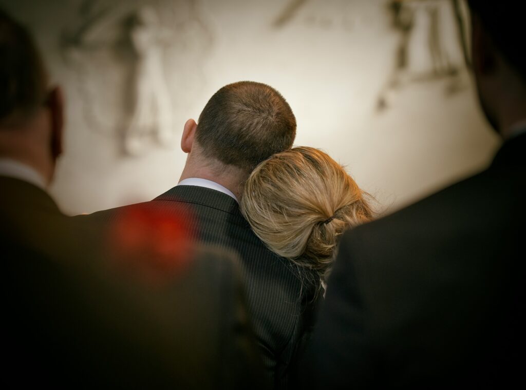 A man and woman in formal attire, their heads leaning together in shared grief, are surrounded by others against a blurred monochromatic backdrop.