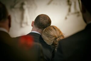 A man and woman in formal attire, their heads leaning together in shared grief, are surrounded by others against a blurred monochromatic backdrop.