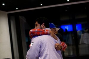 In a dimly lit room, two people, showcasing their human skills of connection and empathy, share an embrace; one wears a striped shirt, while the other sports a plaid shirt with a cap.