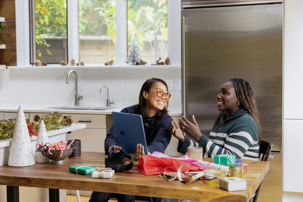 Two people sit at a kitchen table with a laptop and various holiday decorations, including gift boxes and wrapping materials. They appear to be smiling and talking, blending holiday cheer with productivity as they plan the perfect festive season together.