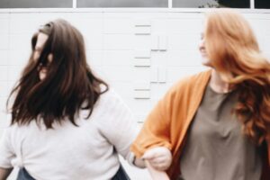 Two people, one with long brown hair and the other with long red hair, are walking hand in hand confidently past a white brick wall, embodying strength and solidarity in a world striving to combat sexual harassment.