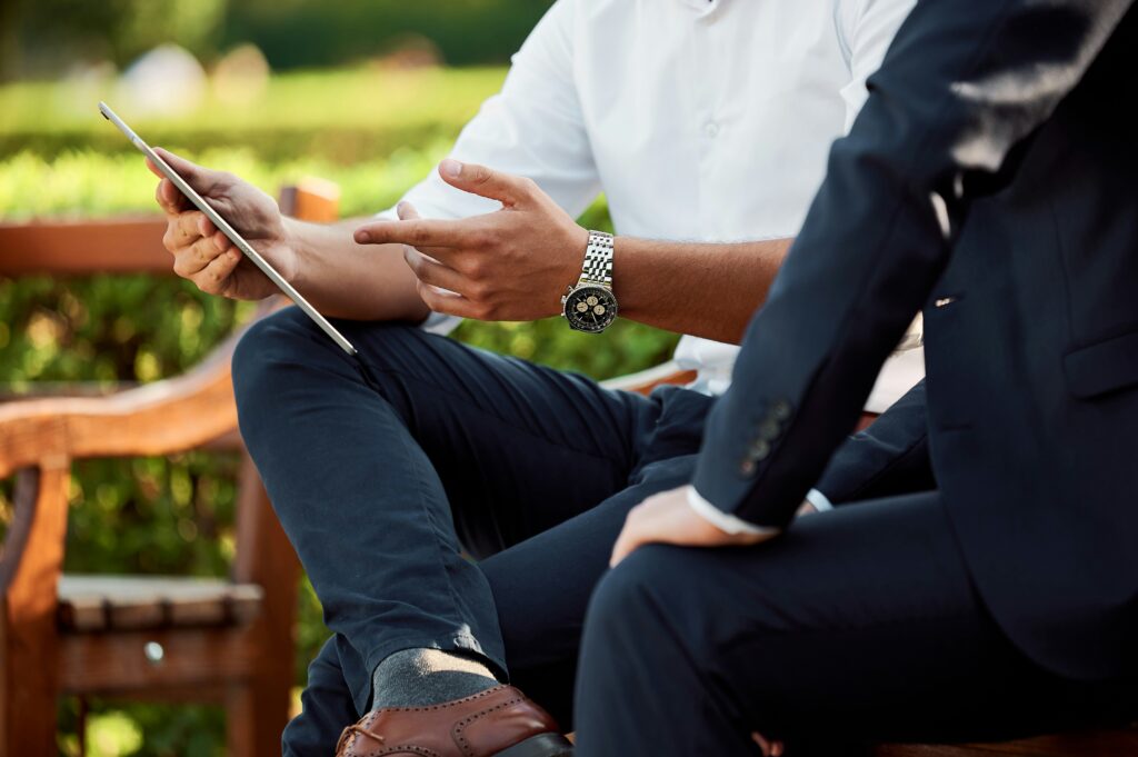 Two individuals, one in a white shirt, comfortably seated outdoors, sharing a tablet as they discuss their budget-friendly plans.